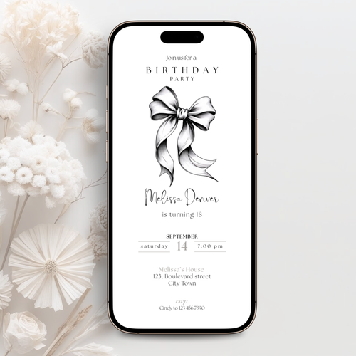 A smartphone centered on a light neutral background, displaying a birthday party invitation design with a large bow illustration and text. White dried flowers are arranged around the phone as styling props.