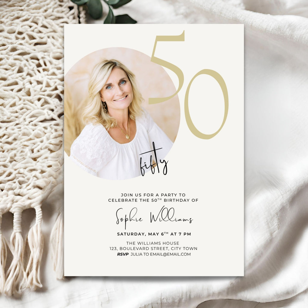 A 50th birthday invitation card styled on a neutral fabric background with a cream macrame runner. The card features a photo of a smiling blonde woman, elegant gold '50' numerals, and black script and serif text for the event details.