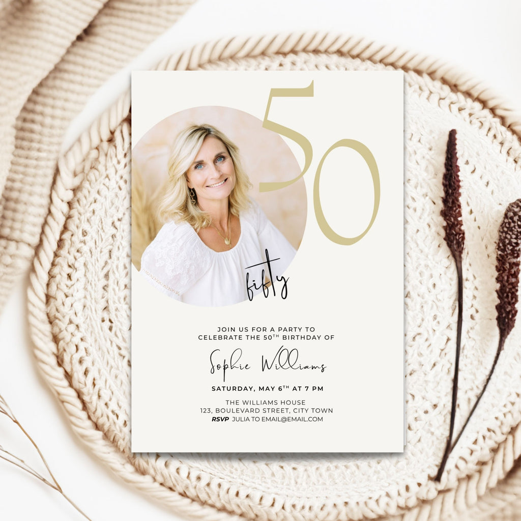 A 50th birthday invitation card styled on a woven beige mat with dried botanicals. The card features a photo of a smiling blonde woman, elegant gold '50' typography, and black script and serif text for the event details.