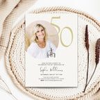 A 50th birthday invitation card styled on a woven beige mat with dried botanicals. The card features a photo of a smiling blonde woman, elegant gold '50' typography, and black script and serif text for the event details.