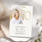 A 50th birthday invitation card styled on a white scalloped plate with soft beige fabric and small white flowers. The card features a photo of a smiling blonde woman, large gold '50' text, and black script and serif text for the event details.