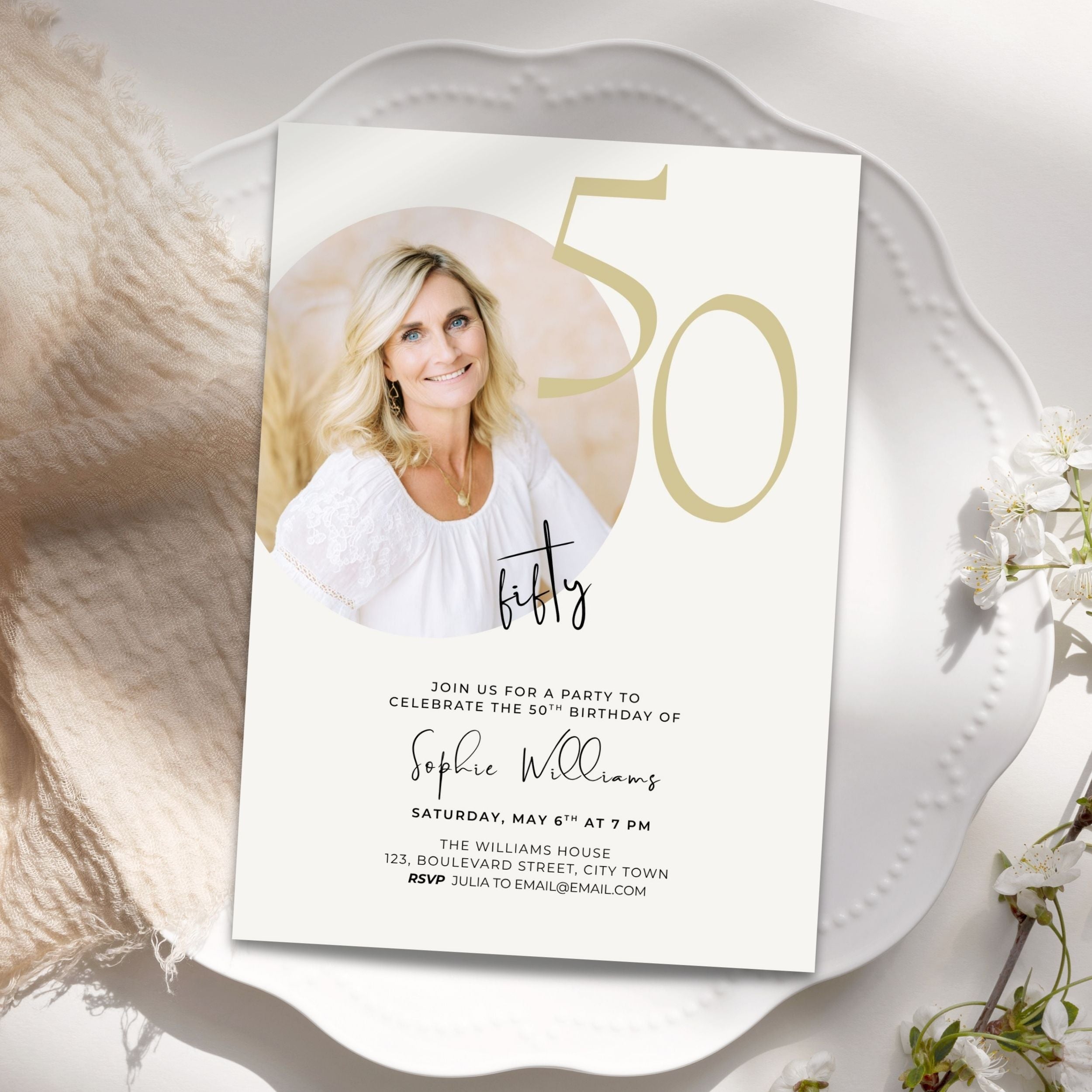 A 50th birthday invitation card styled on a white scalloped plate with soft beige fabric and small white flowers. The card features a photo of a smiling blonde woman, large gold '50' text, and black script and serif text for the event details.