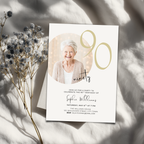 Invitation card for a 90th birthday party with a photo of an elderly woman, placed on a light surface with dried flowers.