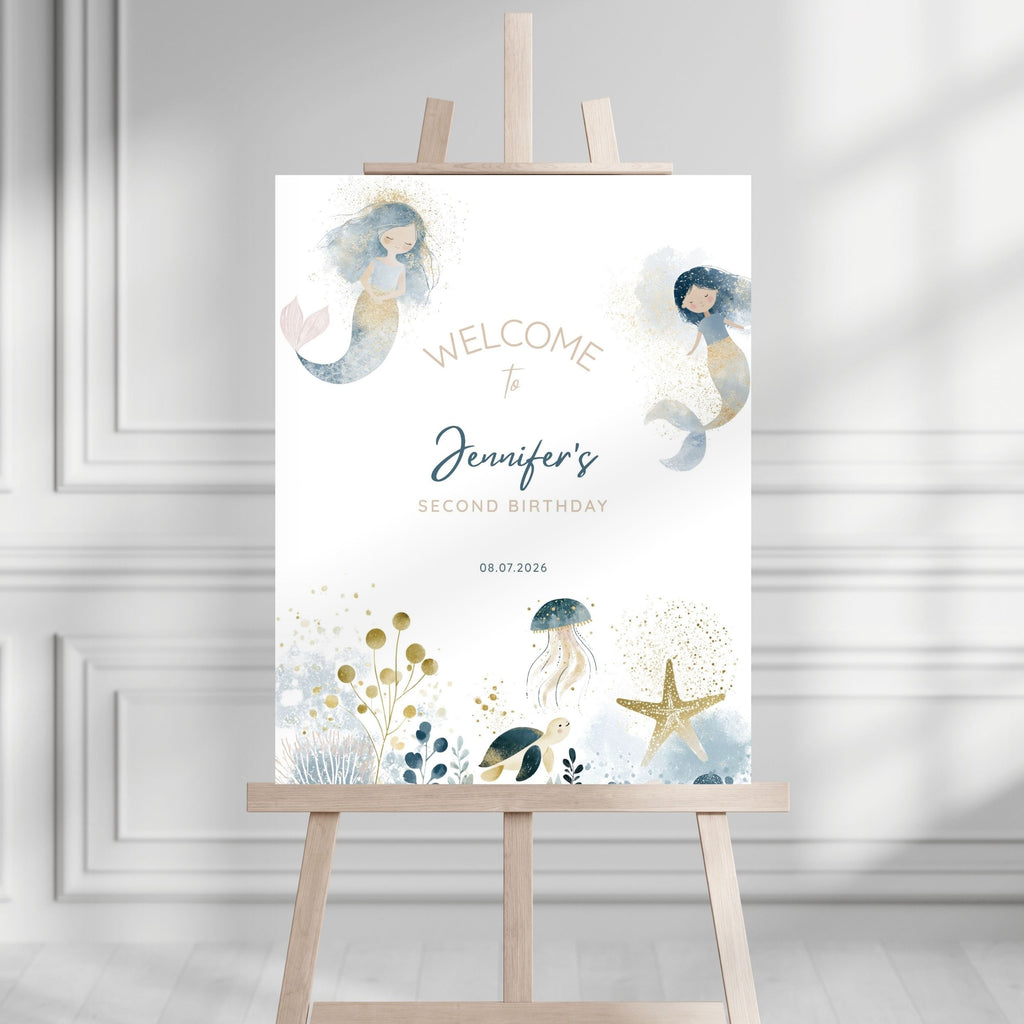 A personalized mermaid-themed second birthday welcome sign displayed on a wooden easel in a bright, modern room with white paneled walls. The sign features watercolor illustrations of two mermaids, a jellyfish, a starfish, and sea plants in soft blue, gold, and gray tones, with customizable text reading 'Welcome to Jennifer's Second Birthday' and the date '08.