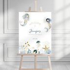 A personalized mermaid-themed second birthday welcome sign displayed on a wooden easel in a bright, modern room with white paneled walls. The sign features watercolor illustrations of two mermaids, a jellyfish, a starfish, and sea plants in soft blue, gold, and gray tones, with customizable text reading 'Welcome to Jennifer's Second Birthday' and the date '08.