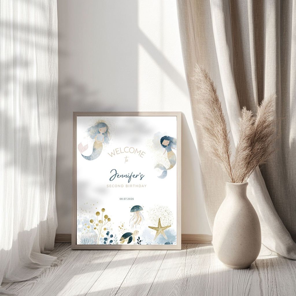 A framed personalized mermaid birthday print leaning against a wall in a bright room with sheer curtains and a vase of pampas grass. The print reads 'Welcome to Jennifer's Second Birthday' with watercolor mermaids and sea-themed illustrations.