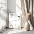 A framed personalized mermaid birthday print leaning against a wall in a bright room with sheer curtains and a vase of pampas grass. The print reads 'Welcome to Jennifer's Second Birthday' with watercolor mermaids and sea-themed illustrations.