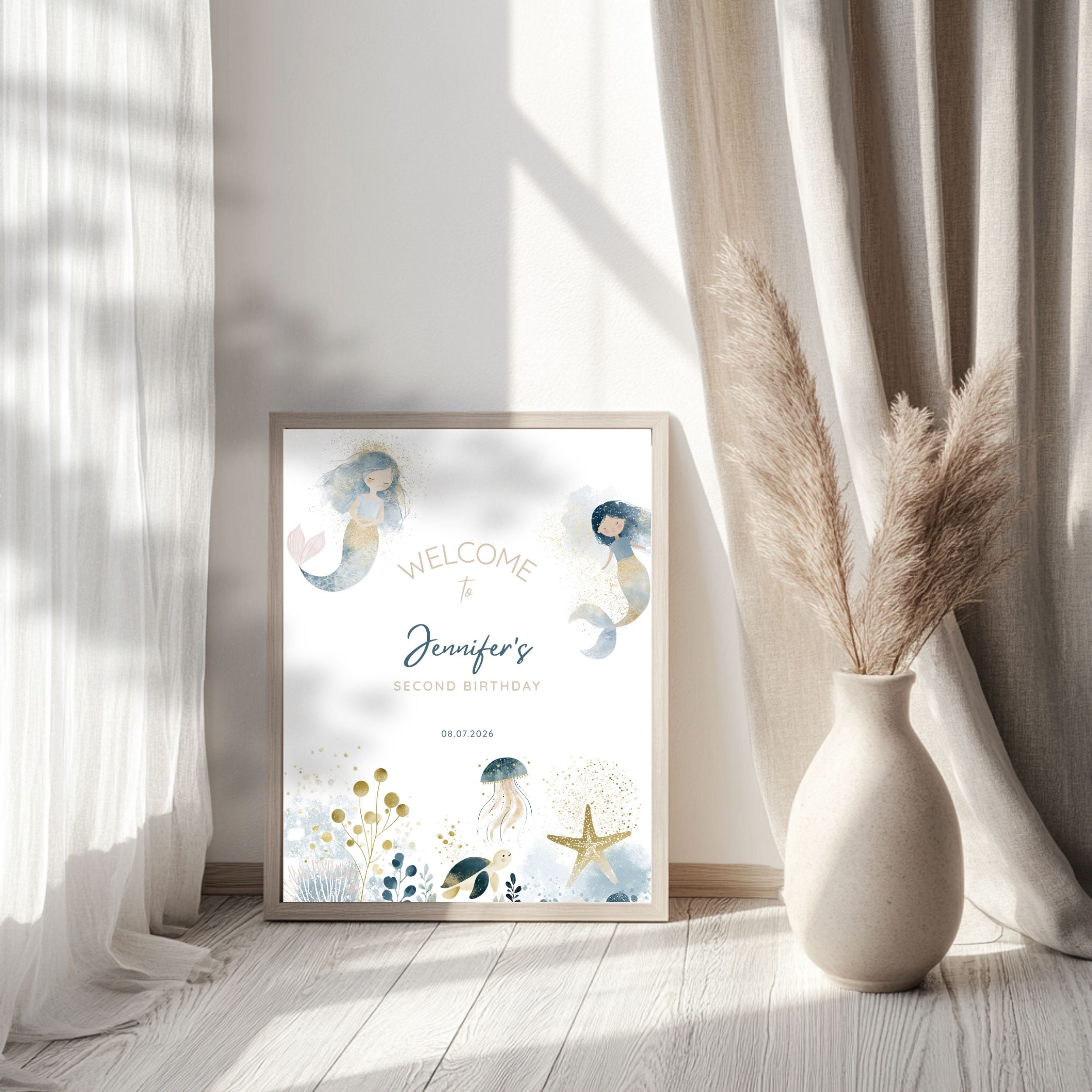 A framed personalized mermaid birthday print leaning against a wall in a bright room with sheer curtains and a vase of pampas grass. The print reads 'Welcome to Jennifer's Second Birthday' with watercolor mermaids and sea-themed illustrations.