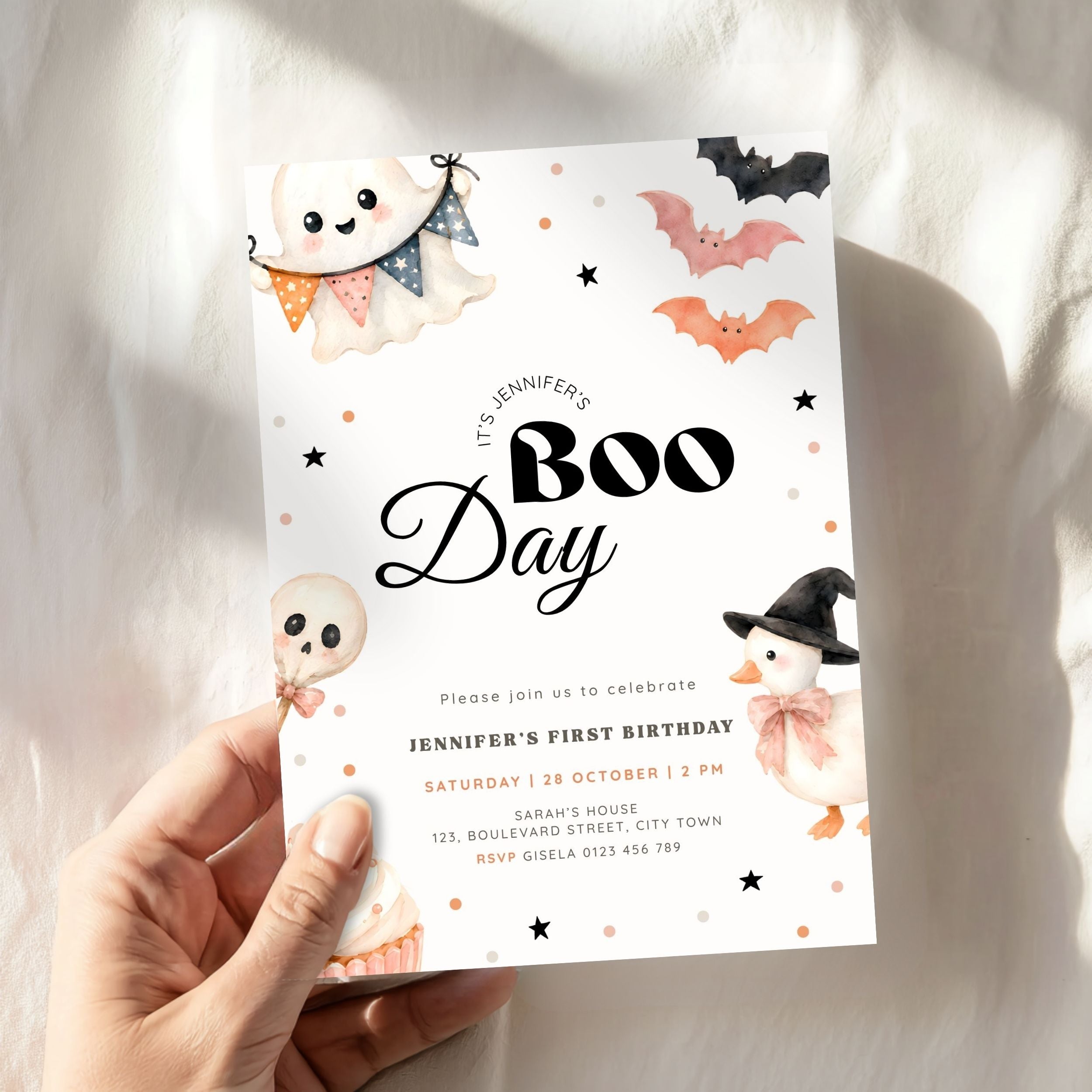 A hand holds a Halloween-themed first birthday invitation card featuring cute ghost, bat, and witch illustrations on a white background with warm sunlight and soft shadows.