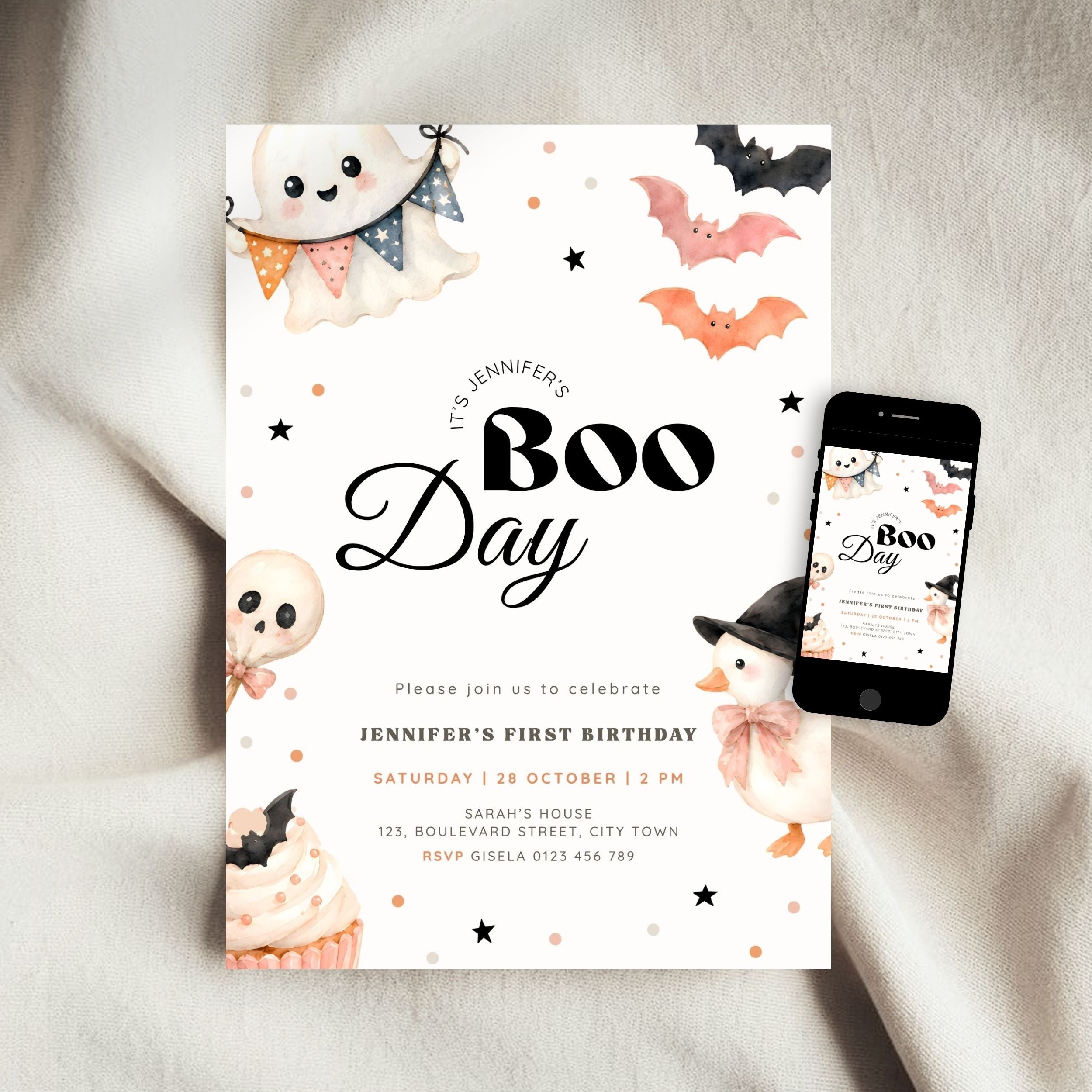 A Halloween-themed first birthday invitation card is styled on a soft beige fabric background next to a smartphone showing the matching digital design. The card features cute watercolor illustrations of a ghost, bat, skull, cupcake, and a witch, with the text 'It's Jennifer's Boo Day'.