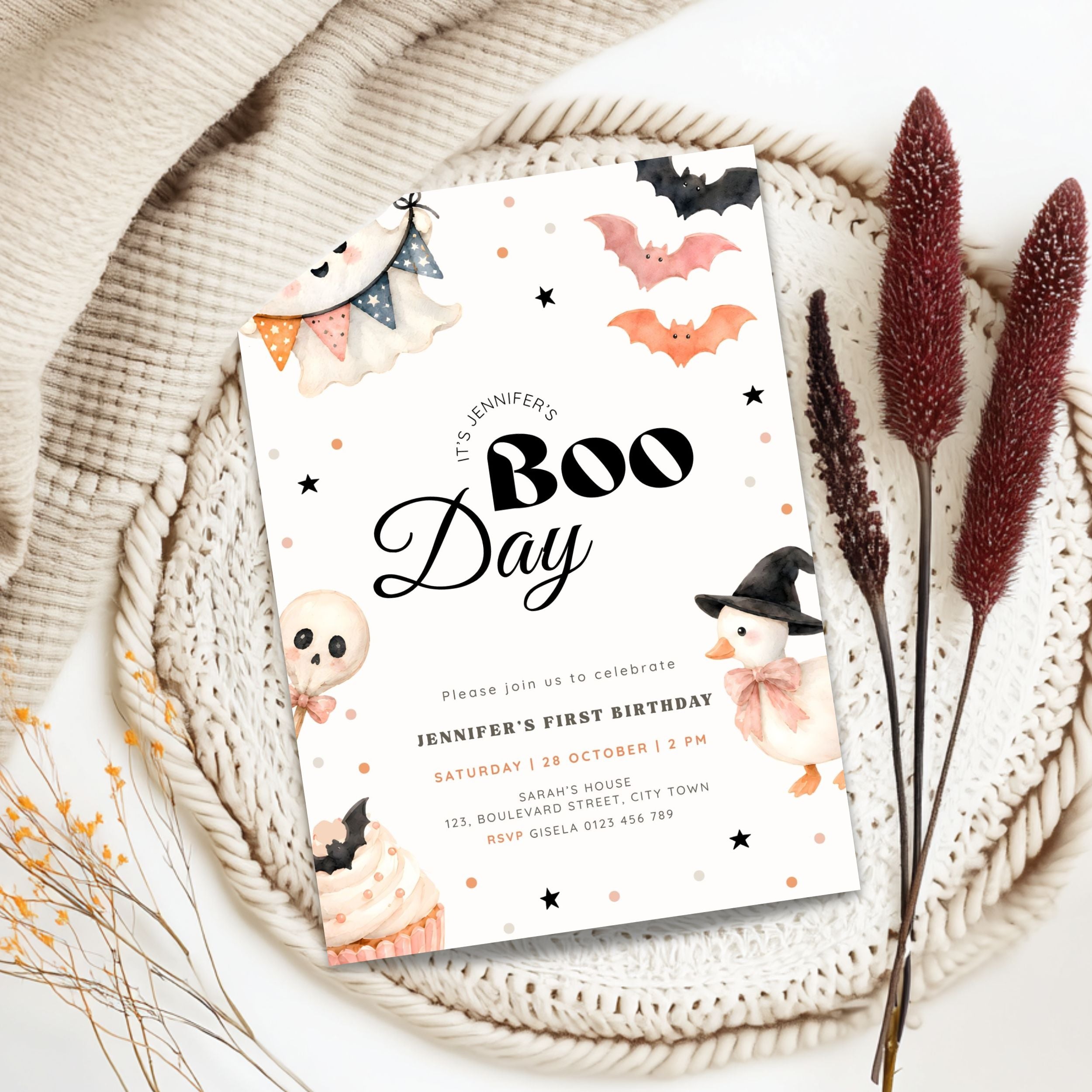 A Halloween-themed birthday invitation card styled in a flat lay on a beige woven placemat with dried decorative grass and a cream knit blanket. The card features cute illustrations of bats, a skull, a ghost, a pumpkin, and a witch, with the text 'It's Jennifer's Boo Day'.