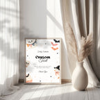 A framed custom Halloween greeting card with cute watercolor illustrations of a ghost, cupcakes, bats, and a witch, styled in a bright room next to a vase of pampas grass.