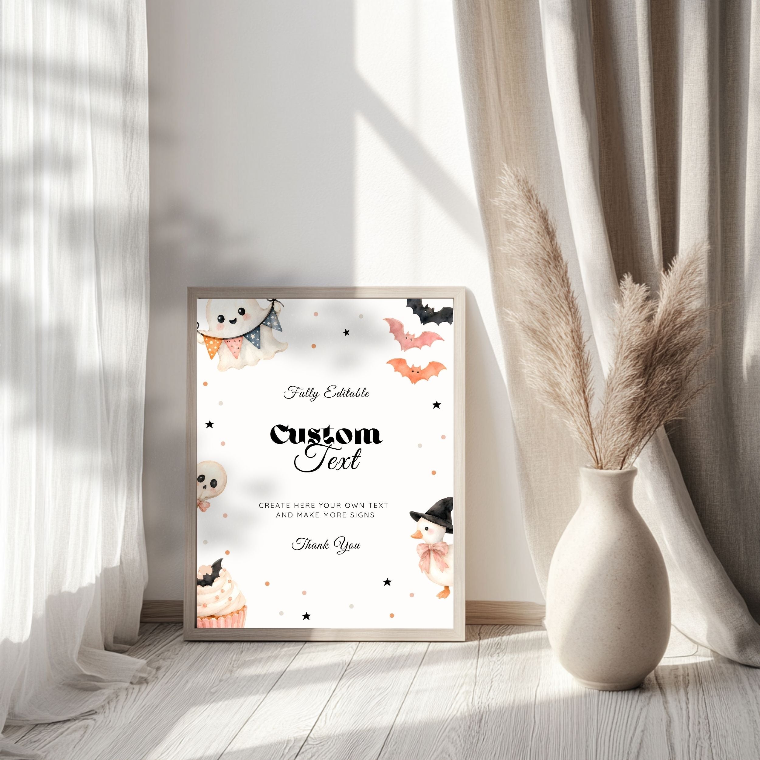 A framed custom Halloween greeting card with cute watercolor illustrations of a ghost, cupcakes, bats, and a witch, styled in a bright room next to a vase of pampas grass.