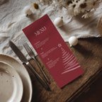 A burgundy Christmas-themed menu card with white text and a tree illustration, styled on a wooden table with a plate, fork, knife, and white decorative ornaments.