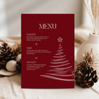 A vertical red menu card with white text reading 'MENU' and holiday dish descriptions, styled on a light surface with pinecones and dried grass in the background.