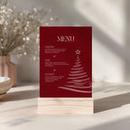 A vertical burgundy christmas menu card with white text and a white Christmas tree illustration, displayed on a light wood stand on a tabletop with soft-focus decorative plants in the background.
