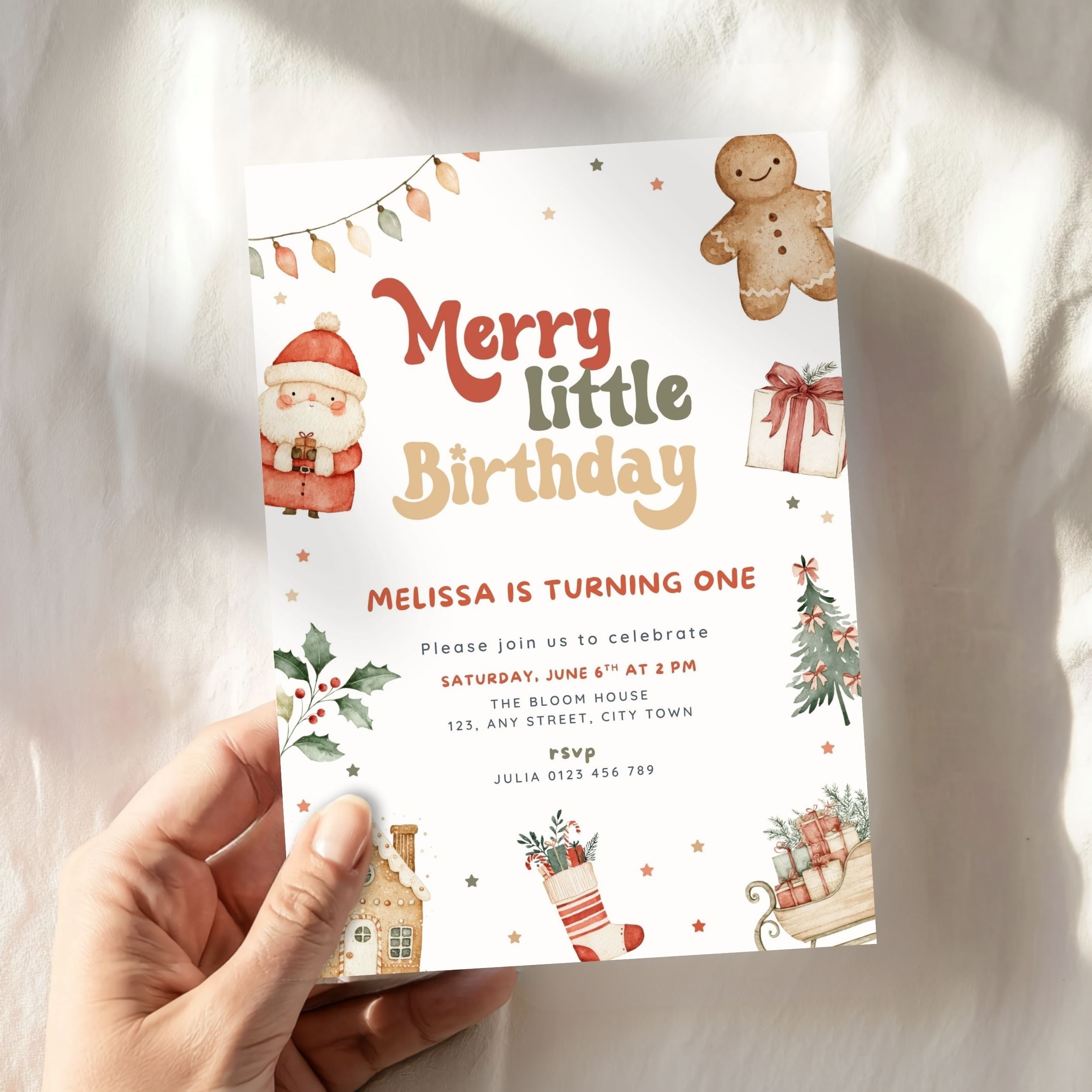 A hand holds a Christmas-themed birthday invitation card with illustrations of a gingerbread man, Santa, gifts, and a stocking, set against a soft white fabric background with warm sunlight.