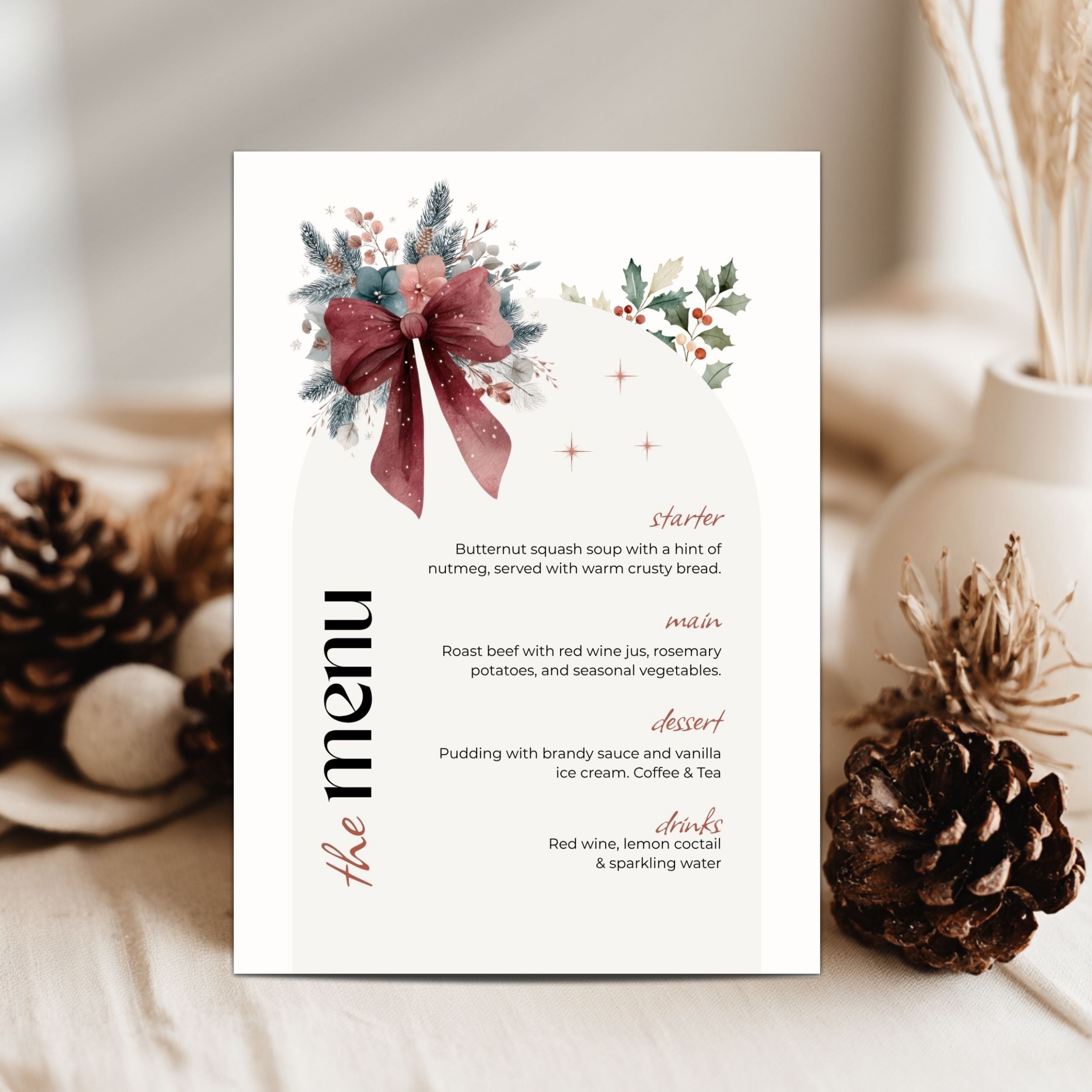 A vertical christmas-style menu card with a white background, watercolor floral and bow illustration at the top, and black and dusty rose text listing course names and items. The card is photographed on a soft neutral surface with pinecones and dried stems around it.