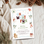 A festive Christmas-themed birthday invitation card styled in a flat-lay arrangement on a cozy beige knit blanket, surrounded by pine needles, pinecones, and a gingerbread cookie.