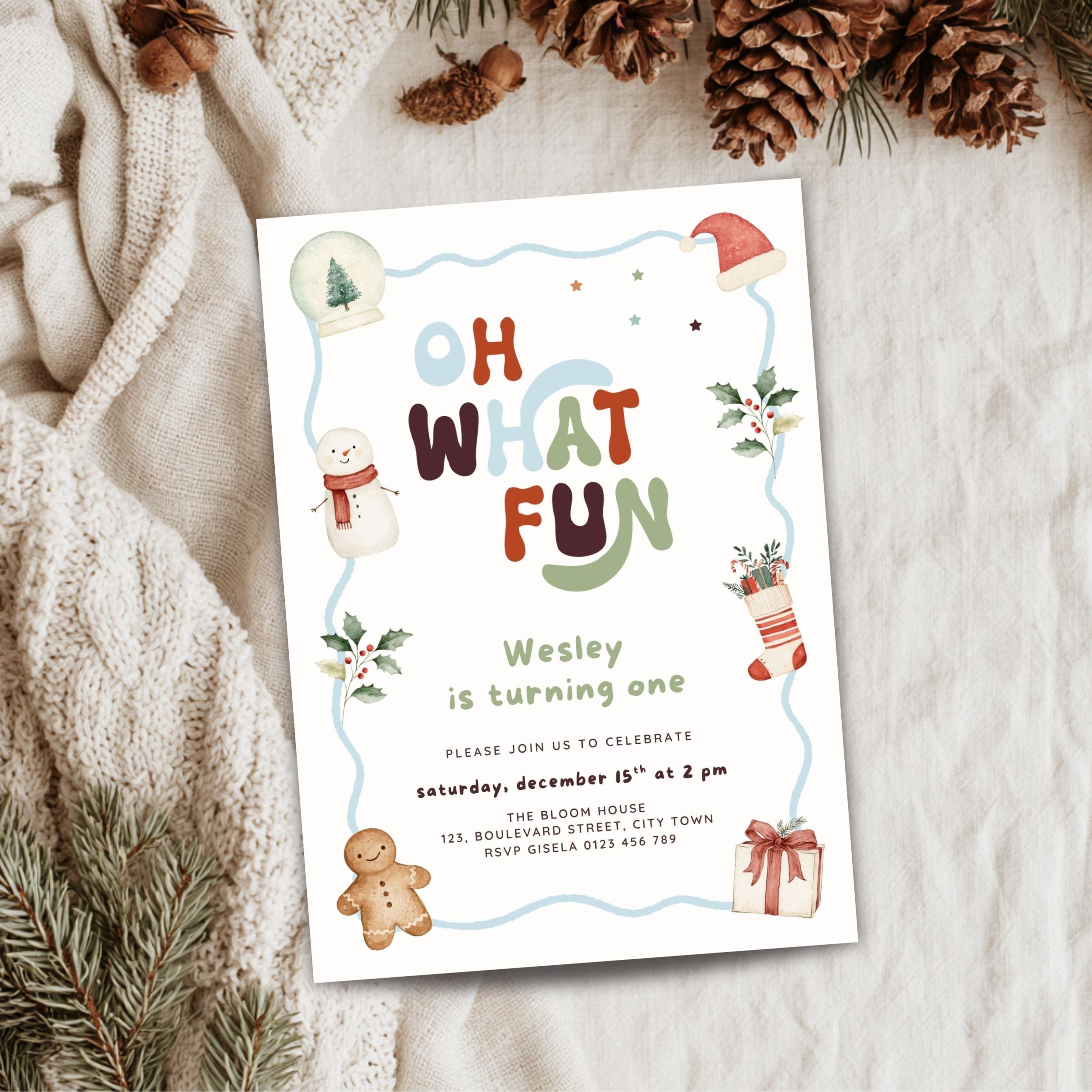 A festive Christmas-themed birthday invitation card styled in a flat-lay arrangement on a cozy beige knit blanket, surrounded by pine needles, pinecones, and a gingerbread cookie.