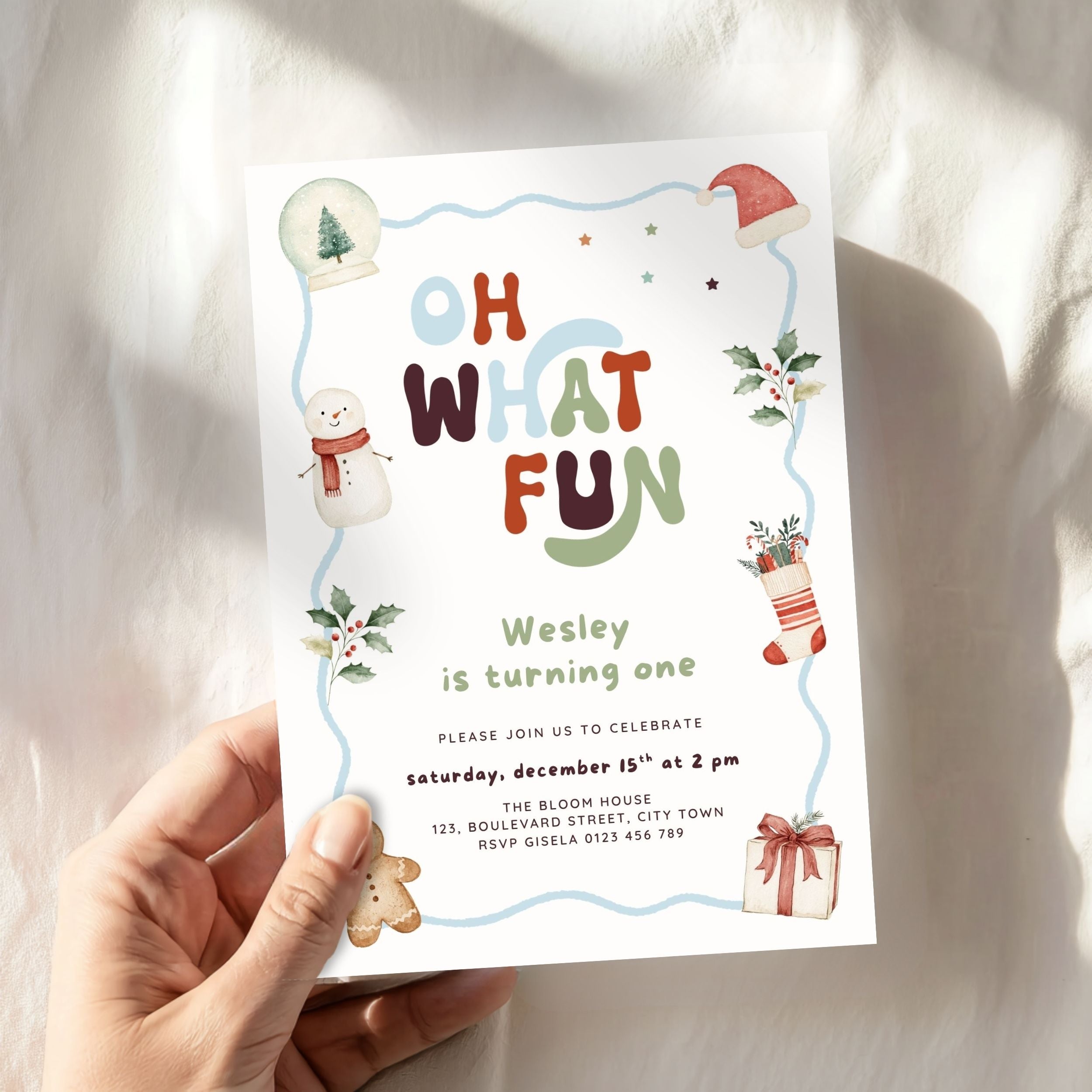 A hand holds a festive Christmas-themed birthday invitation card against a softly lit white fabric background. The card features colorful playful text reading 'OH WHAT FUN' surrounded by holiday illustrations including a snowman, Santa hat, snow globe, holly, stocking, gingerbread cookie, and gift box.