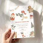 A hand holding a Christmas-themed birthday invitation card with festive illustrations of Santa, a gingerbread man, a stocking, and holiday decorations on a soft white fabric background.