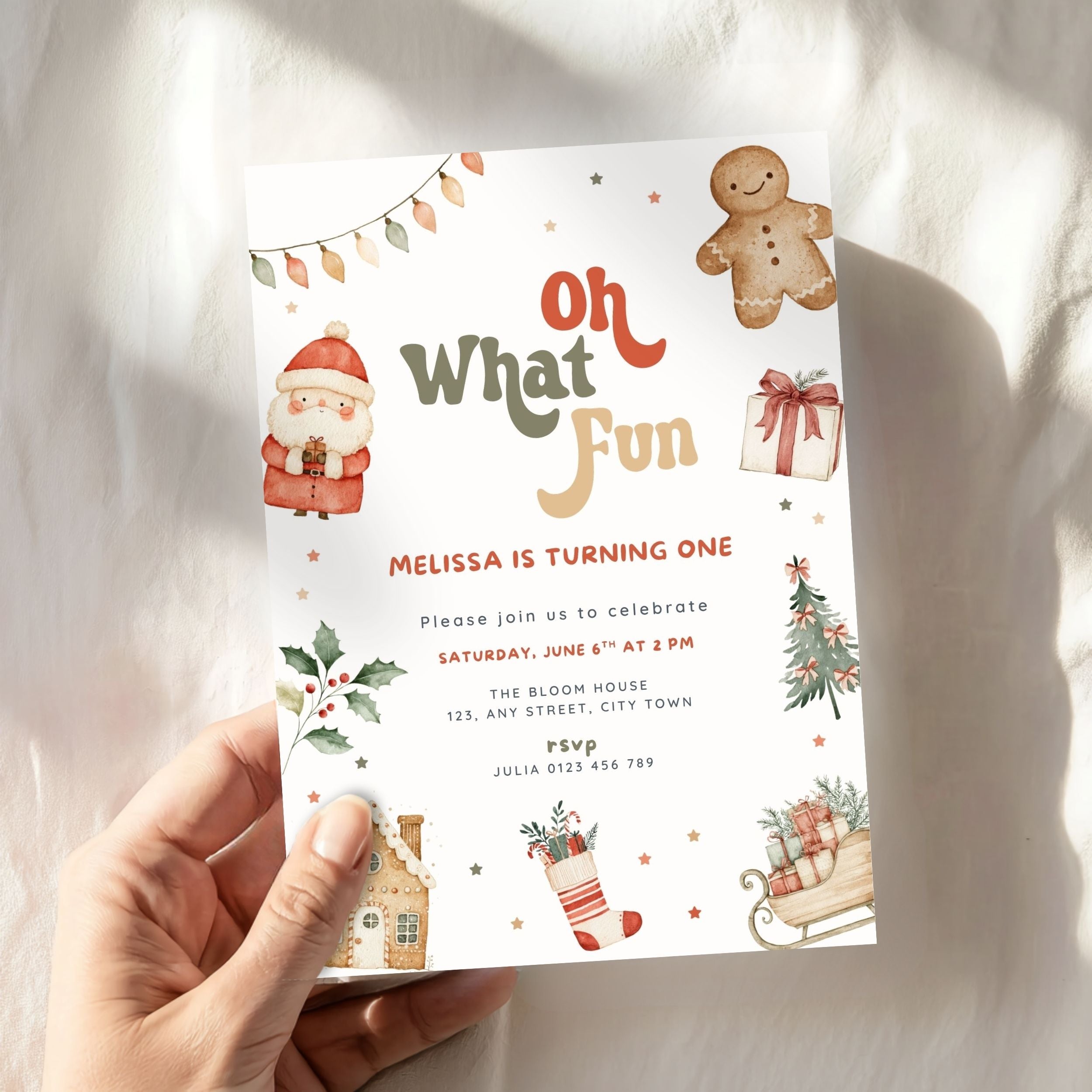 A hand holding a Christmas-themed birthday invitation card with festive illustrations of Santa, a gingerbread man, a stocking, and holiday decorations on a soft white fabric background.
