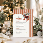 A festive holiday menu card with orange and white vertical stripes, a red bow illustration, and elegant script text, displayed on a table with blurred Christmas decorations and a tree in the background.