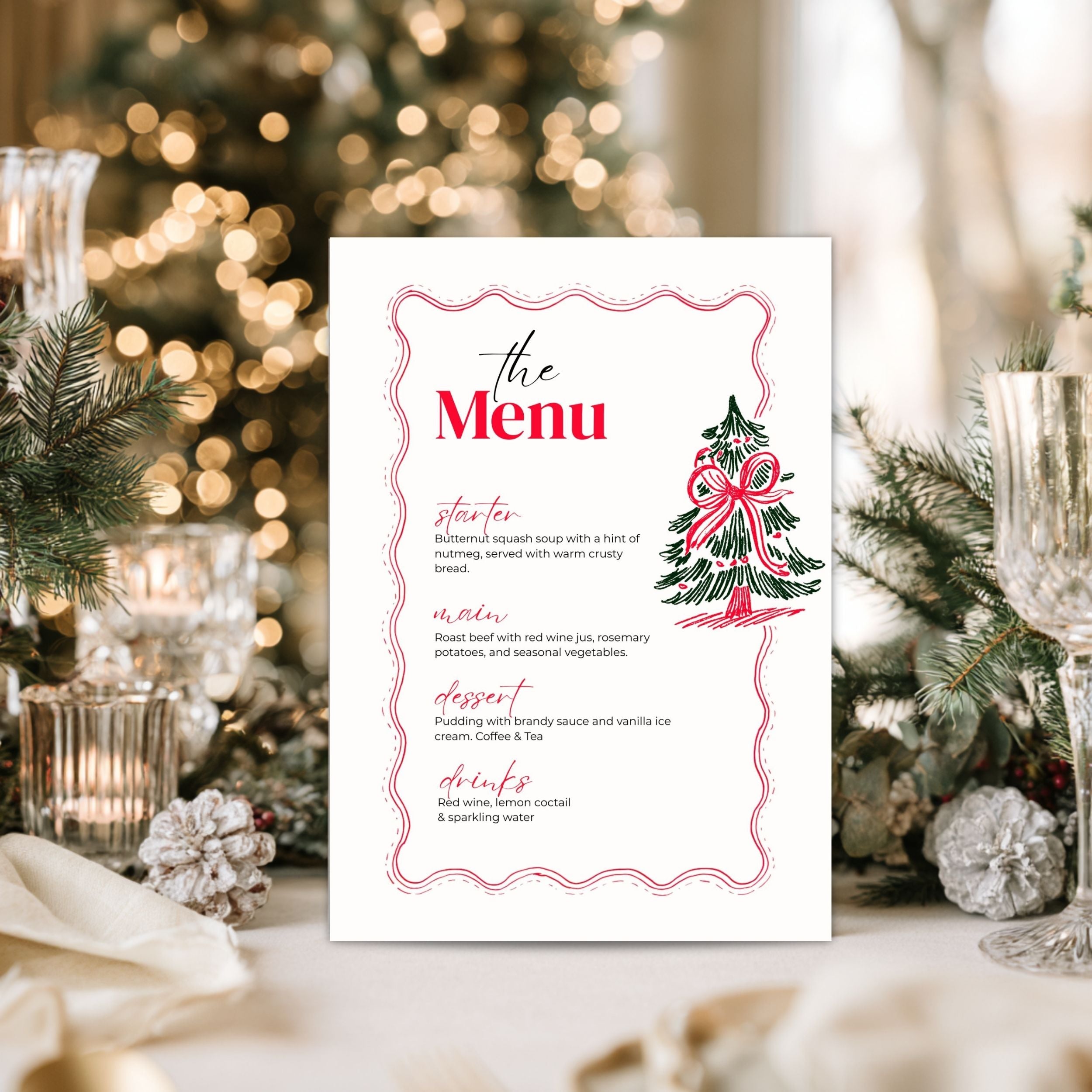 A printed menu card with a pink decorative border, red title text reading 'The Menu,' and a small illustrated Christmas tree with a bow, photographed standing on a tabletop in a festive holiday setting with blurred lights and greenery in the background.