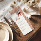 A printed paper menu card styled on a wooden table with a knife, fork, plates, and dried flowers. The menu has rust-orange striped borders, a red bow illustration, and black text reading 'The Menu' with course headings below.