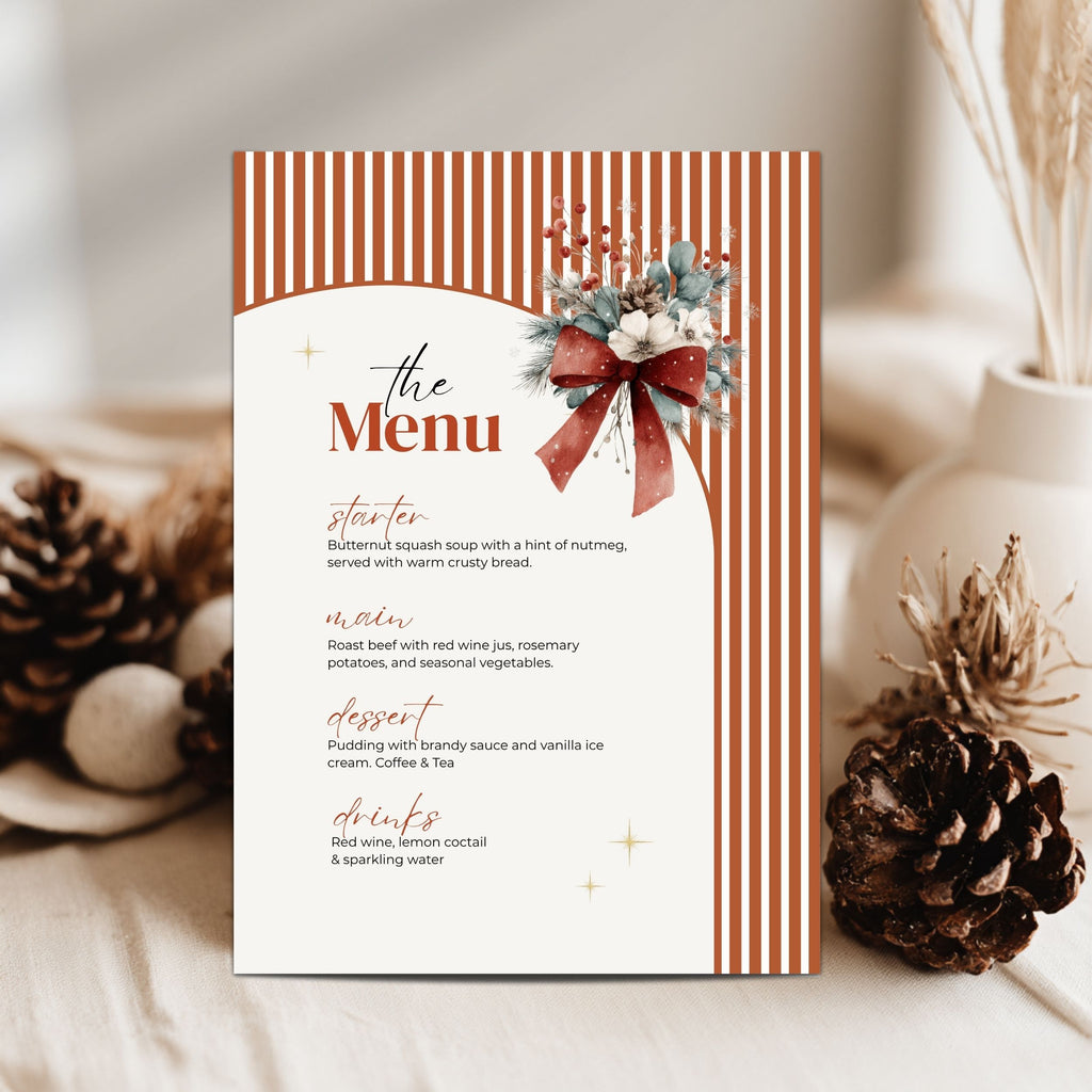 A vertical christmas menu card styled on a neutral fabric surface with pinecones and dried grass. The card has orange and white vertical stripes, a cream center panel with elegant script text, and a decorative floral bow illustration near the top right.