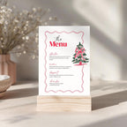 A printed menu card with a red decorative border, red title text reading 'the Menu,' and a small Christmas tree illustration, displayed on a light wood block stand on a tabletop in a softly lit interior setting.