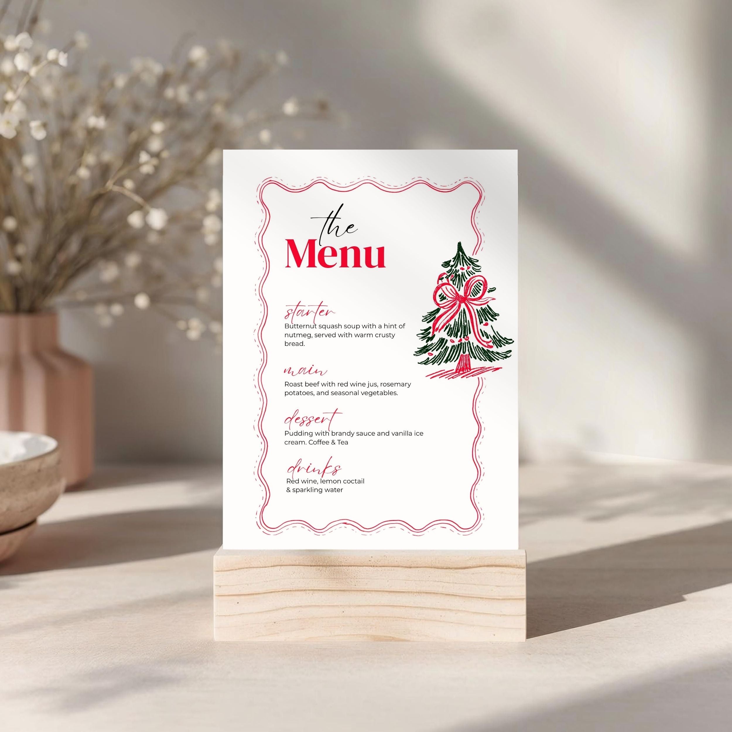 A printed menu card with a red decorative border, red title text reading 'the Menu,' and a small Christmas tree illustration, displayed on a light wood block stand on a tabletop in a softly lit interior setting.