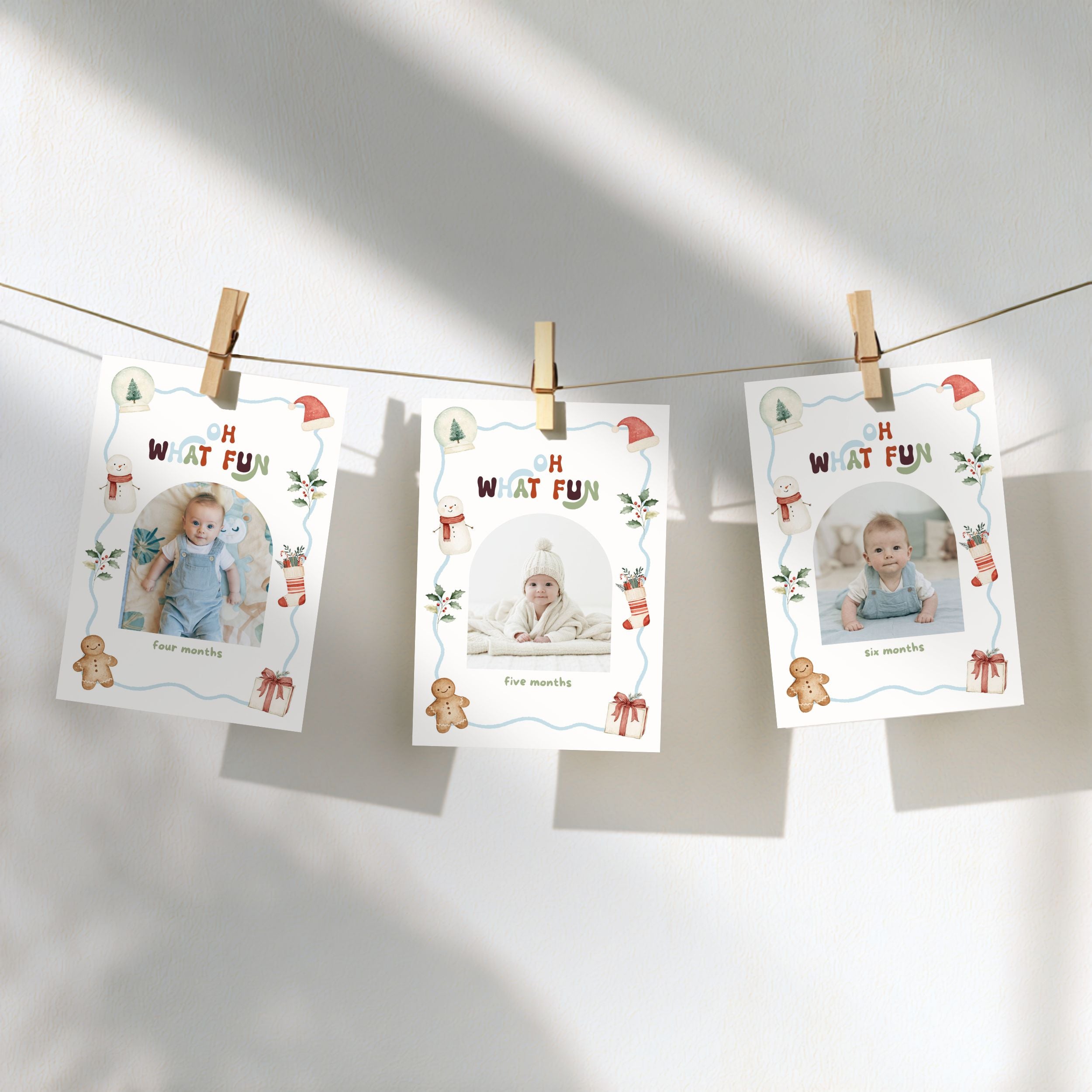 Three printed baby milestone photos hang from a string with wooden clothespins against a white wall. Each card has a holiday-themed border with text such as 'How are you' and placeholders for monthly photos.