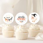 Three Halloween-themed cupcake toppers displayed on top of frosted cupcakes. The toppers feature cute illustrations: a smiling ghost holding a pennant banner, the words 'spooky one' in orange script with a small pumpkin, and a witch duck wearing a black hat and pink bow.