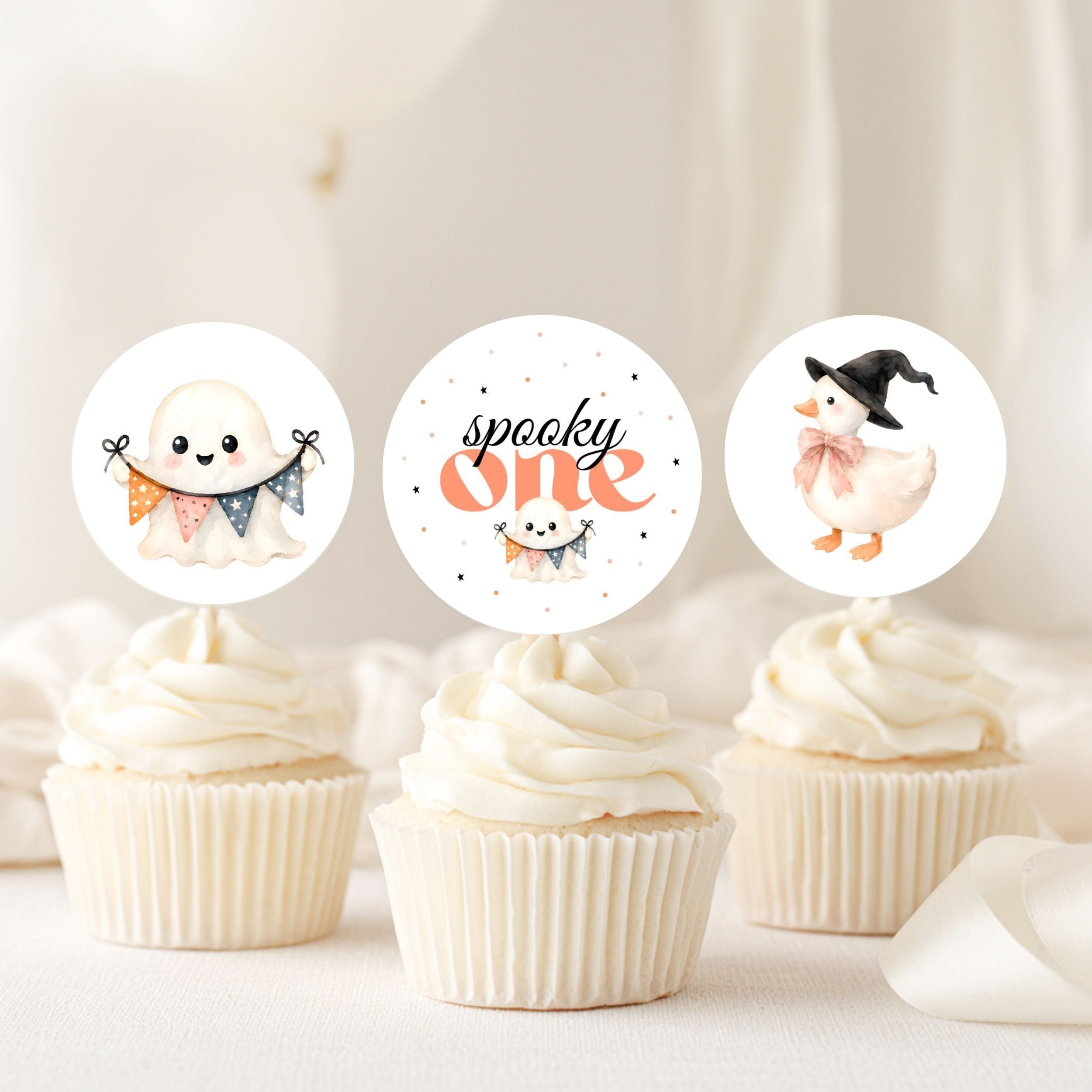 Three Halloween-themed cupcake toppers displayed on top of frosted cupcakes. The toppers feature cute illustrations: a smiling ghost holding a pennant banner, the words 'spooky one' in orange script with a small pumpkin, and a witch duck wearing a black hat and pink bow.