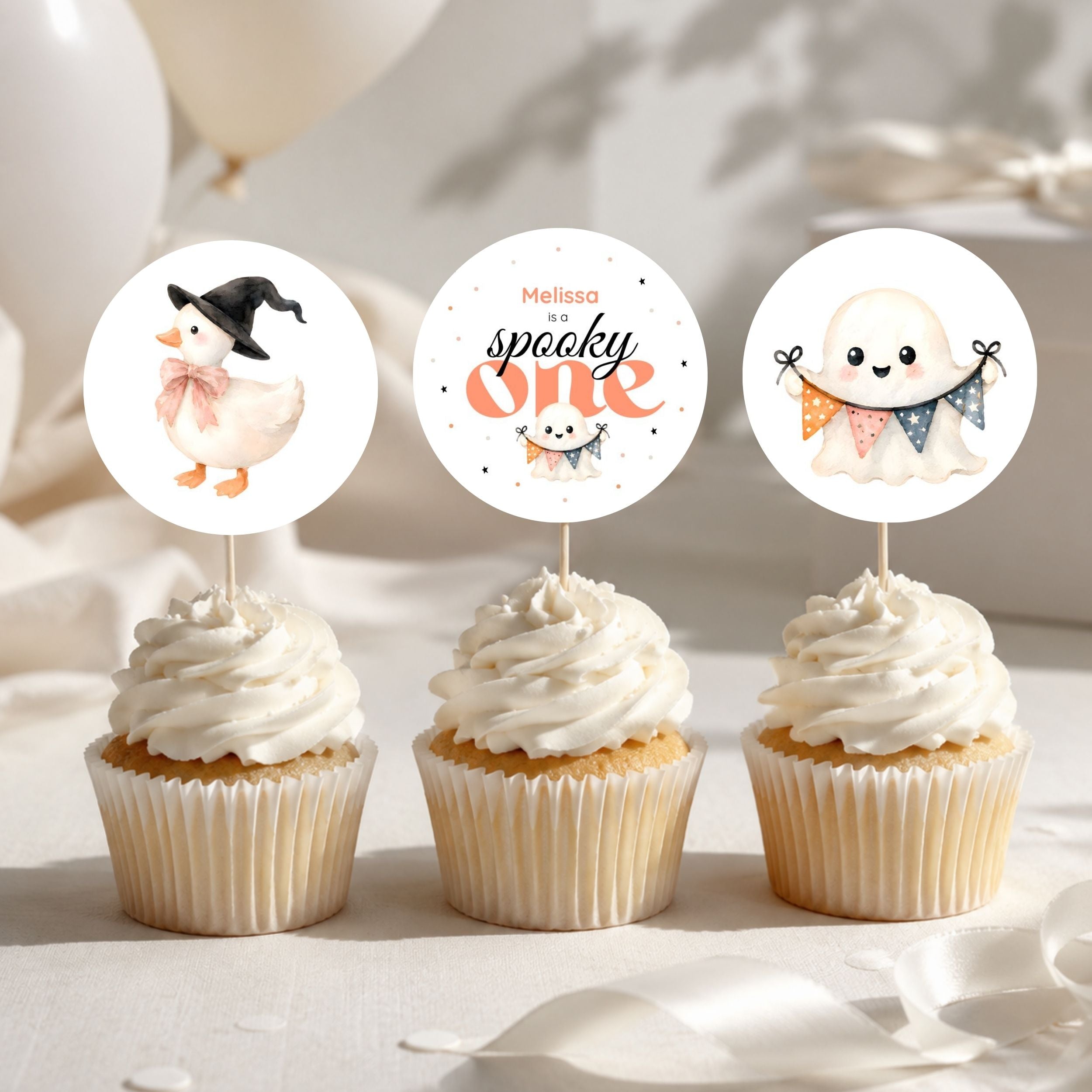 Three cupcakes with round paper topper stickers on a soft white surface. The toppers show Halloween-themed illustrations: a duck in a witch hat, a ghost with colorful flags, and a smiling ghost with the text 'Melissa is spooky one'.