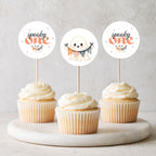 Three frosted cupcakes on a marble tray with round wooden picks featuring cute ghost illustrations and the words 'spooky one' in orange and black.