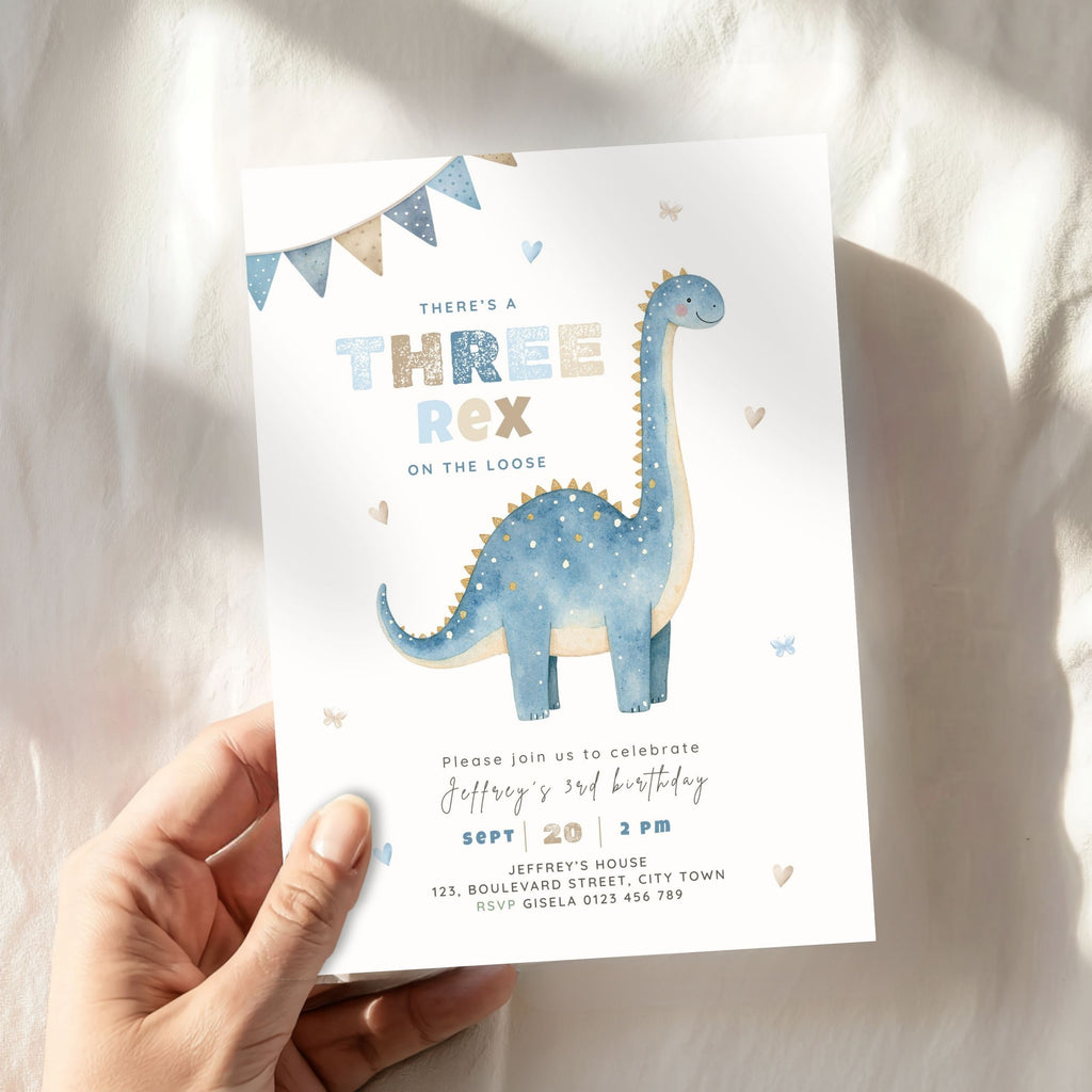 A hand holds a children's dino 3rd birthday invitation card featuring a watercolor blue dinosaur on a white background, styled with bunting and heart accents against a softly lit fabric backdrop.