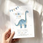 A hand holds a children's dino 3rd birthday invitation card featuring a watercolor blue dinosaur on a white background, styled with bunting and heart accents against a softly lit fabric backdrop.