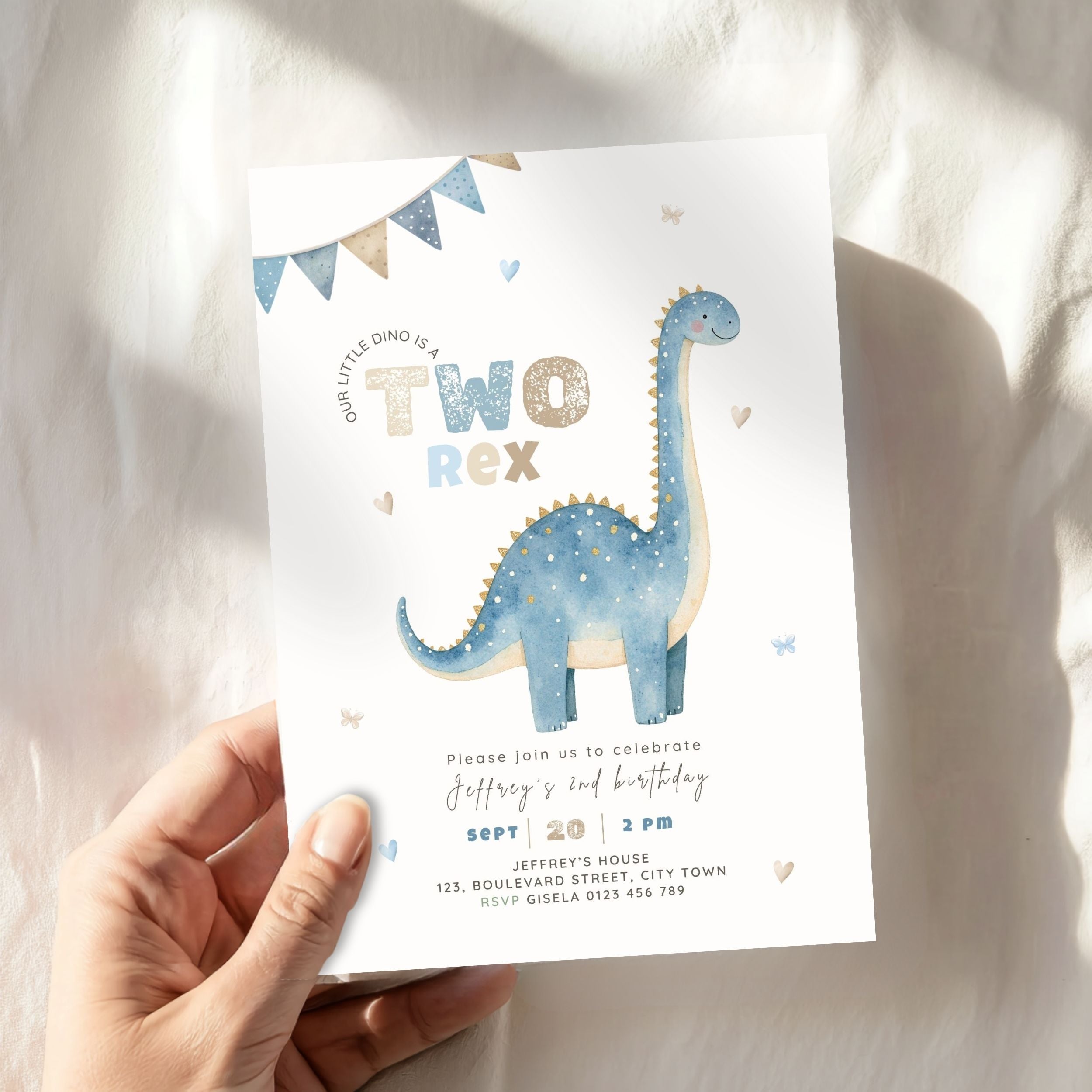 A hand holding a printed dinosaur-themed 2nd birthday invitation card against a softly lit white fabric background. The card features a blue and cream long-neck dinosaur, bunting, and text for a child's second birthday.