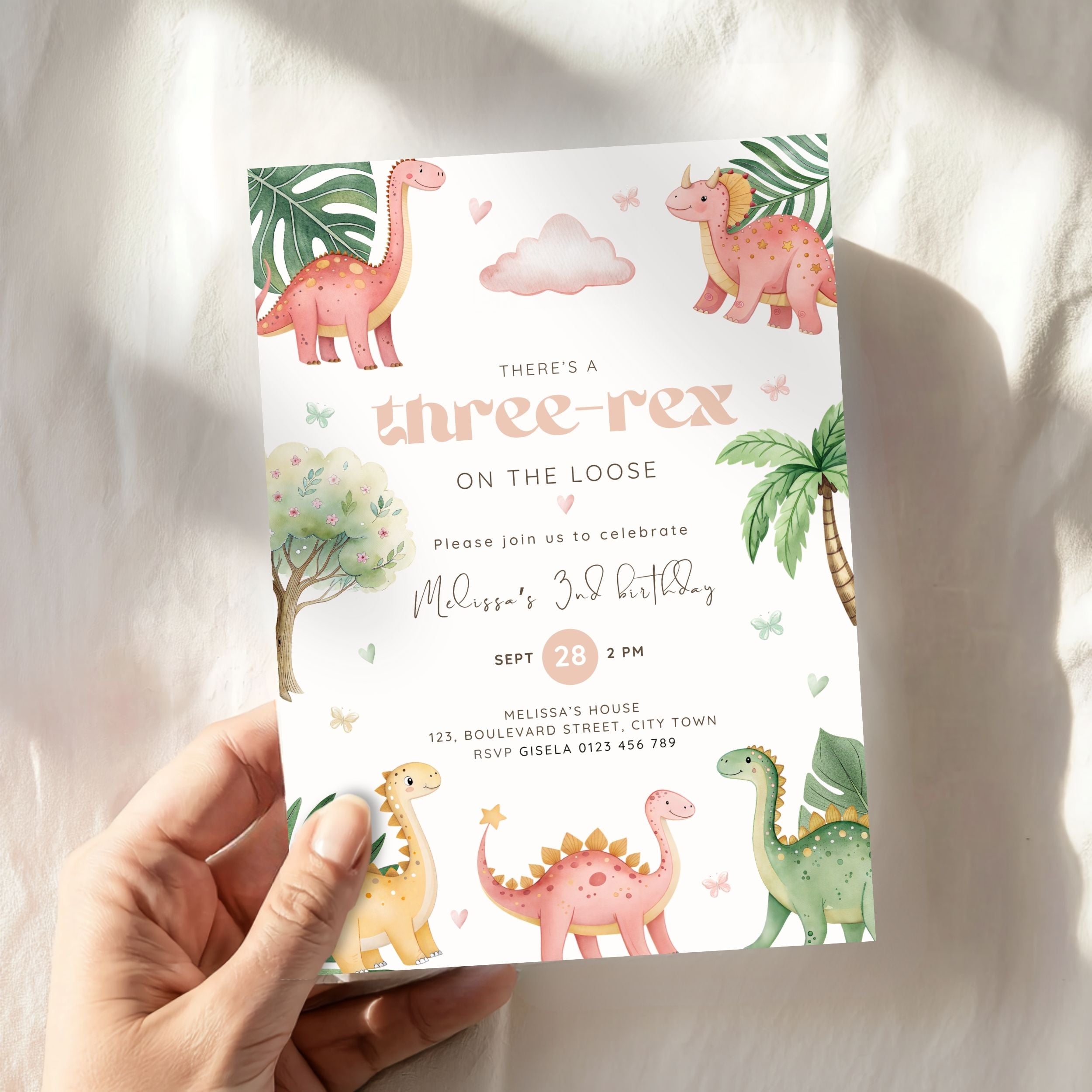 A hand holds a colorful dinosaur-themed 3rd birthday invitation card against a soft white fabric background with natural shadows.