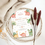 A dinosaur-themed three-rex 3rd birthday invitation card styled in a flat lay with a beige knit placemat, a textured cream knit runner, dried burgundy grass stems, and a soft beige knit blanket.