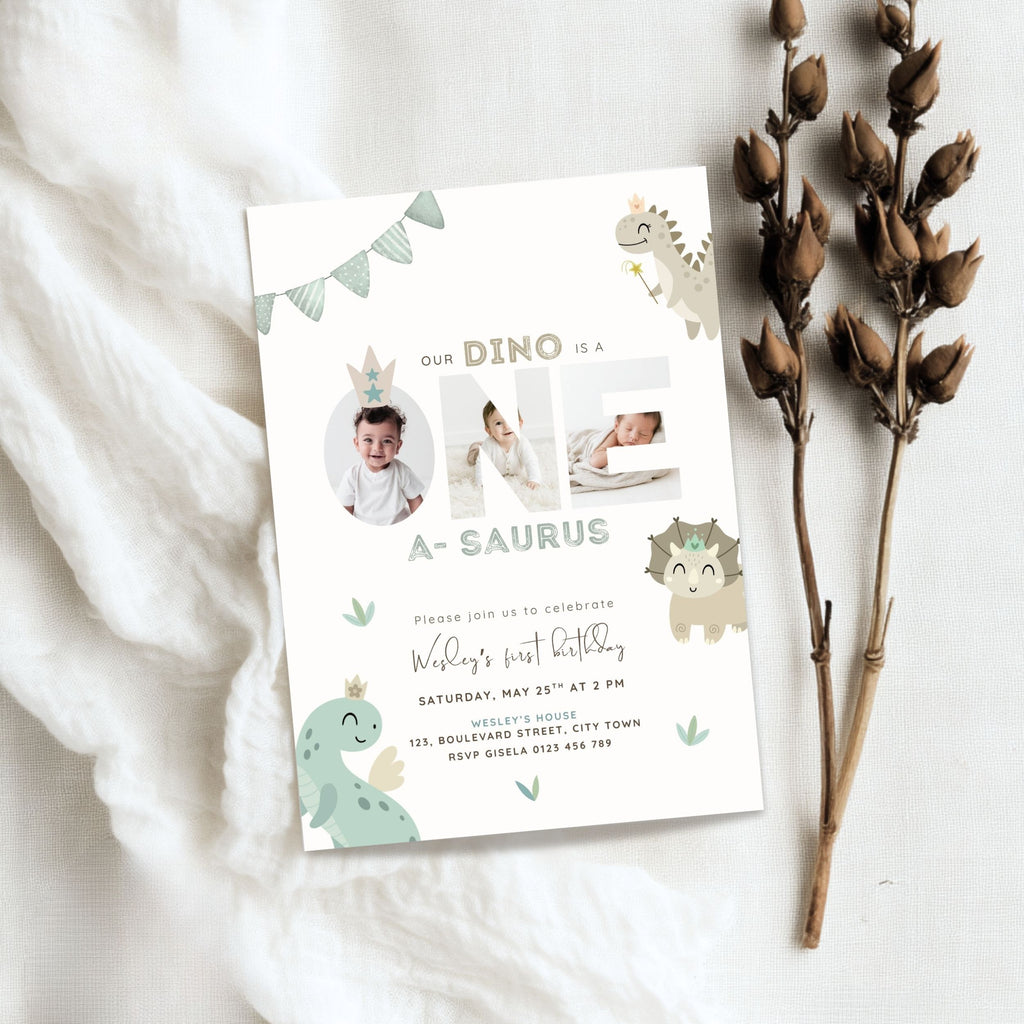 A dinosaur-themed baby first birthday invitation card styled in a flat lay on a white textured fabric background, accompanied by dried brown flower stems. The card features cute cartoon dinosaurs, a bunting banner, and photo slots for the baby.