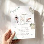 A hand holds a customizable dinosaur-themed first birthday invitation card against a soft, white draped fabric background with warm sunlight shadows.