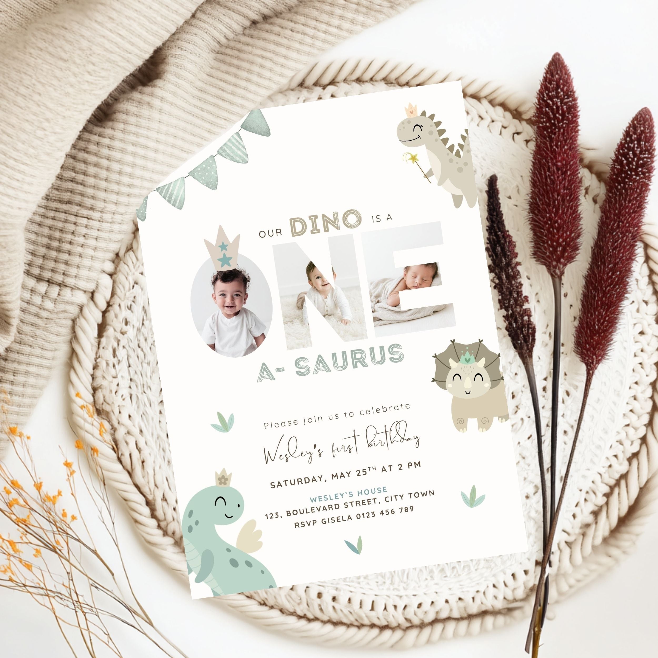 A dinosaur-themed baby first birthday invitation card styled in a flat lay on a beige knit blanket with dried decorative grasses. The white card features cute cartoon dinosaurs, bunting, and placeholder photo slots for a baby.