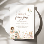 A vertical fairy-themed first birthday party invitation card displayed on a round white plate with soft shadows. The card features elegant script text reading 'Alysia's fairy first birthday party' with watercolor illustrations of fairies, a small cottage, and wildflowers in soft pastel tones.