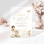 A fairy-themed first birthday invitation card featuring soft pastel illustrations of a dragonfly, a small cottage, and woodland characters on a white background, styled with dried flowers in the background.