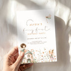 A hand holds a fairy-themed first birthday invitation card with watercolor illustrations of a cottage, flowers, and a dragonfly, set against a softly textured white fabric background with natural shadows.