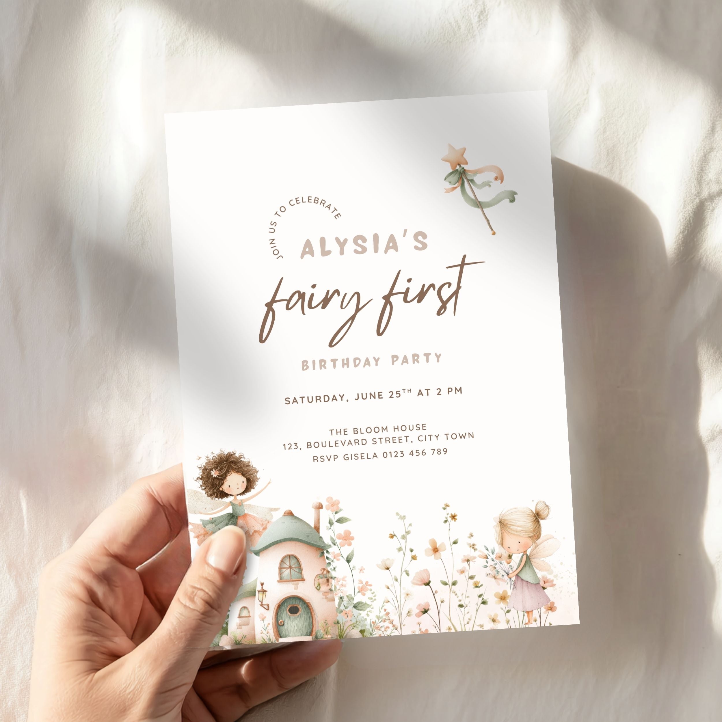 A hand holds a white fairy-themed first birthday invitation card with gold and pink text and pastel illustrations of fairies, flowers, and a small cottage, photographed against a softly lit white fabric background.